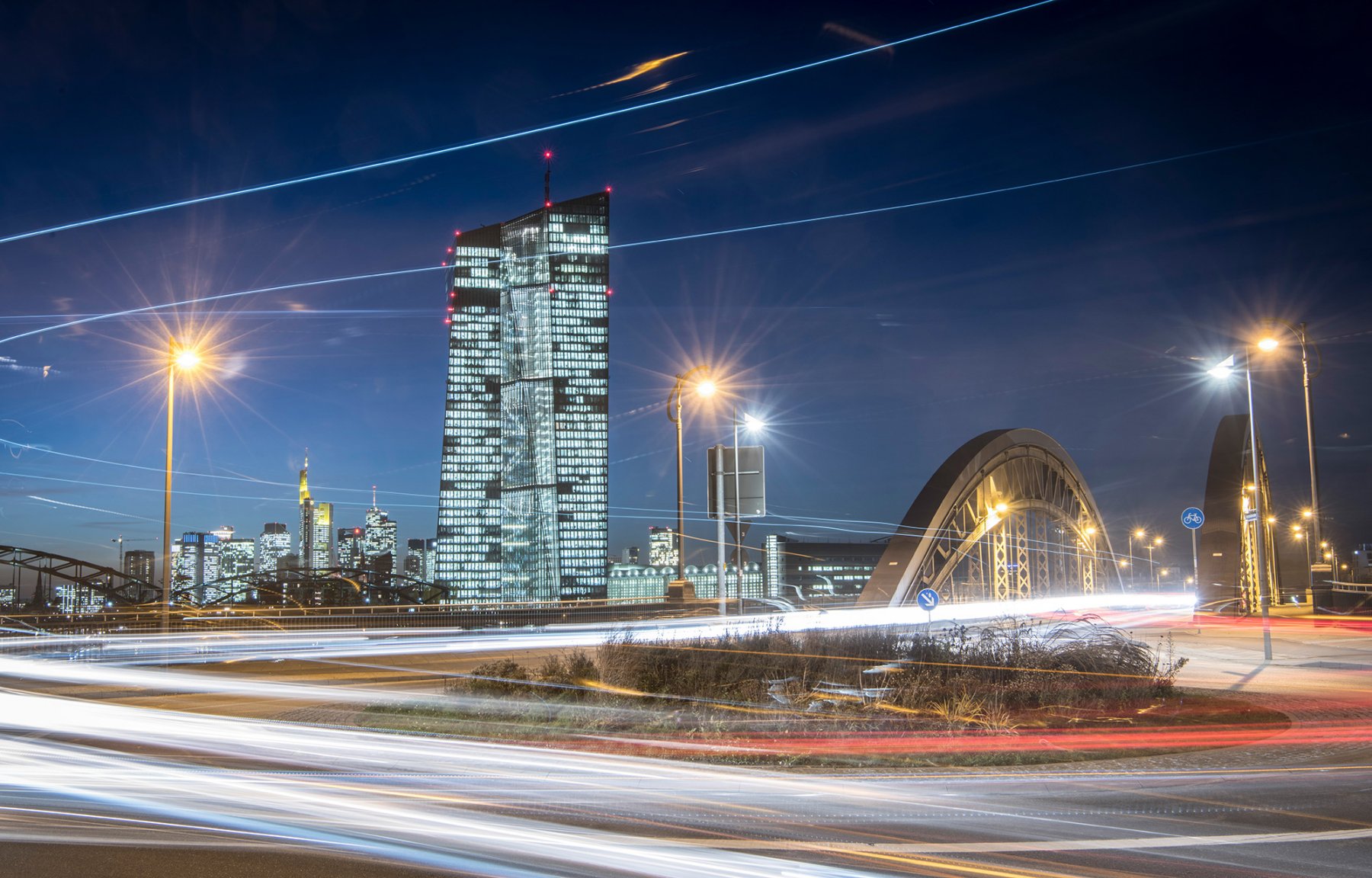 Die Lichter in den Büros der Zentrale der Europäischen Zentralbank leuchten vor der abendlichen Skyline während vorbeifahrende Autos Lichtspuren durch die Dunkelheit ziehen.