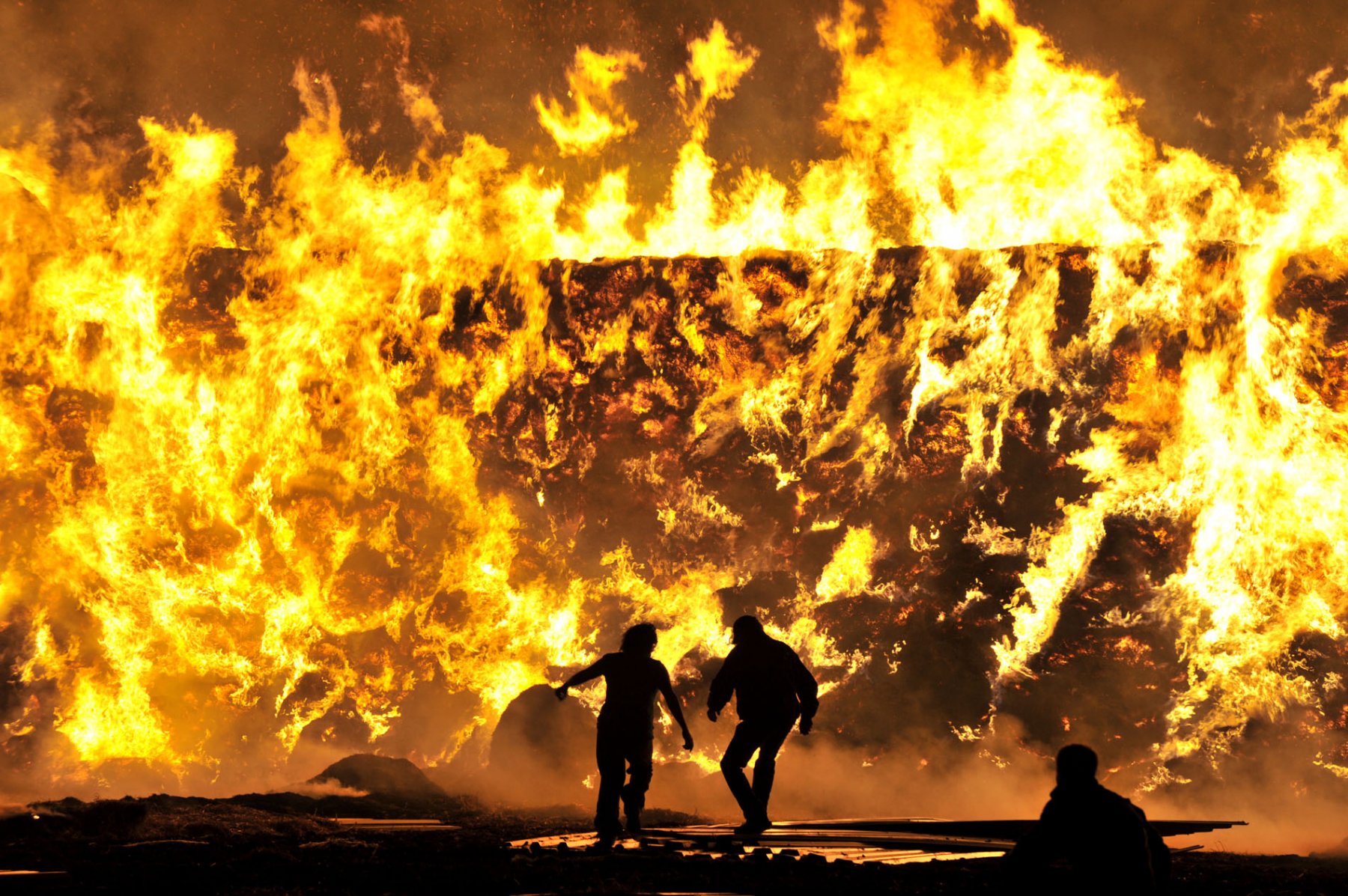 Nur silhouettenhaft zu erkennen sind zwei Menschen, die bei Hattersheim in Hessen vor der Hitze einer lichterloh, brennenden Mauer aus Heuballen davonlaufen. Foto: Boris Roessler/dpa